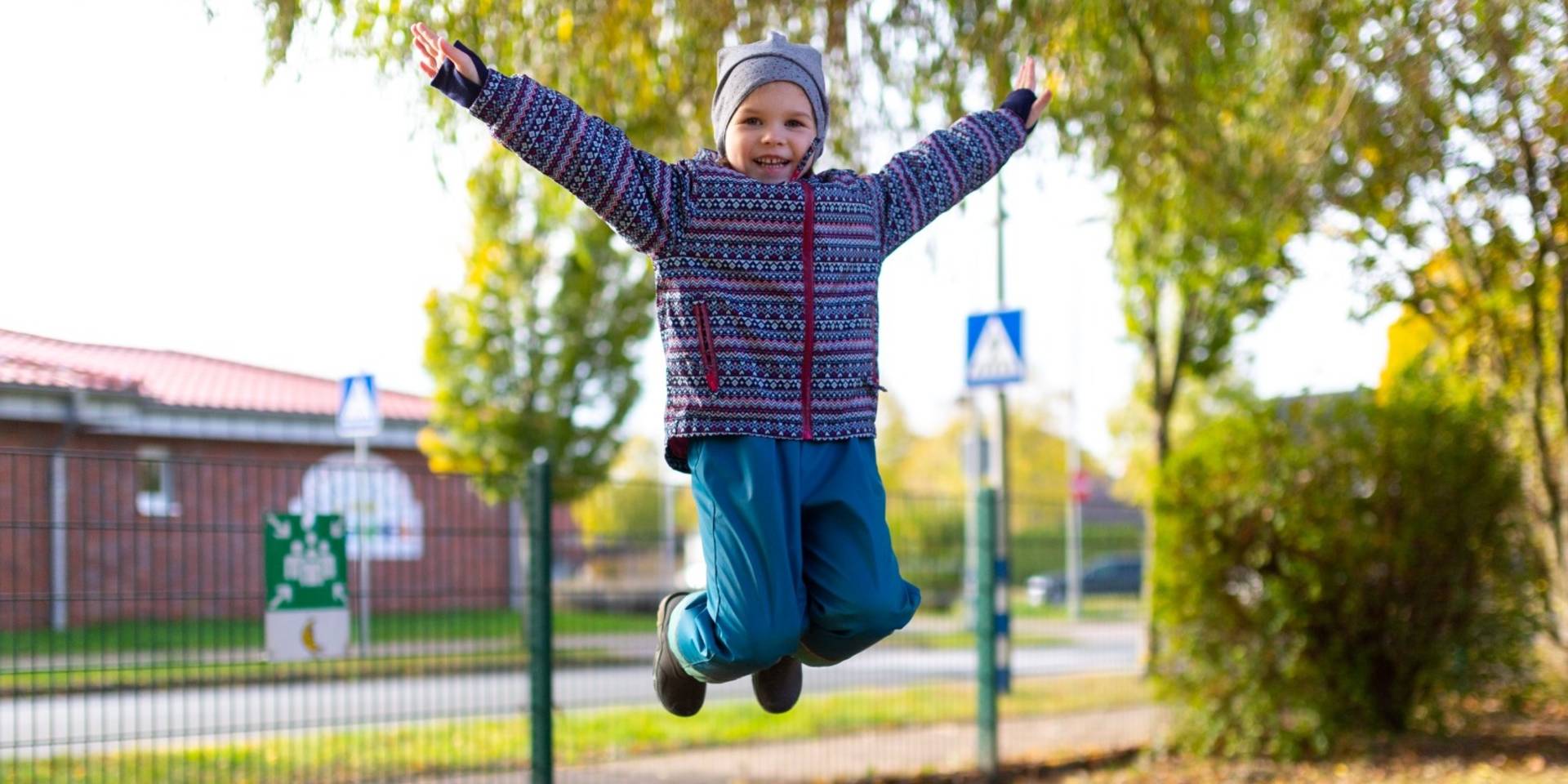 Ein Mädchen springt ganz hoch auf einem Trampolin. Es hat die Knie im Sprung angewinkelt und beide Arme in die Höhe gestreckt.